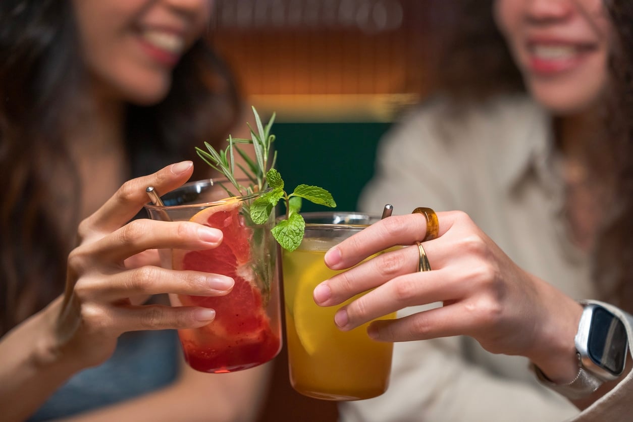 Two young Asian Chinese women, a lesbian couple, toasting colorful cocktails and smiling warmly at each other in a cozy restaurant, celebrating and enjoying the moment, warm lighting and modern interior, Kuala Lumpur, Malaysia